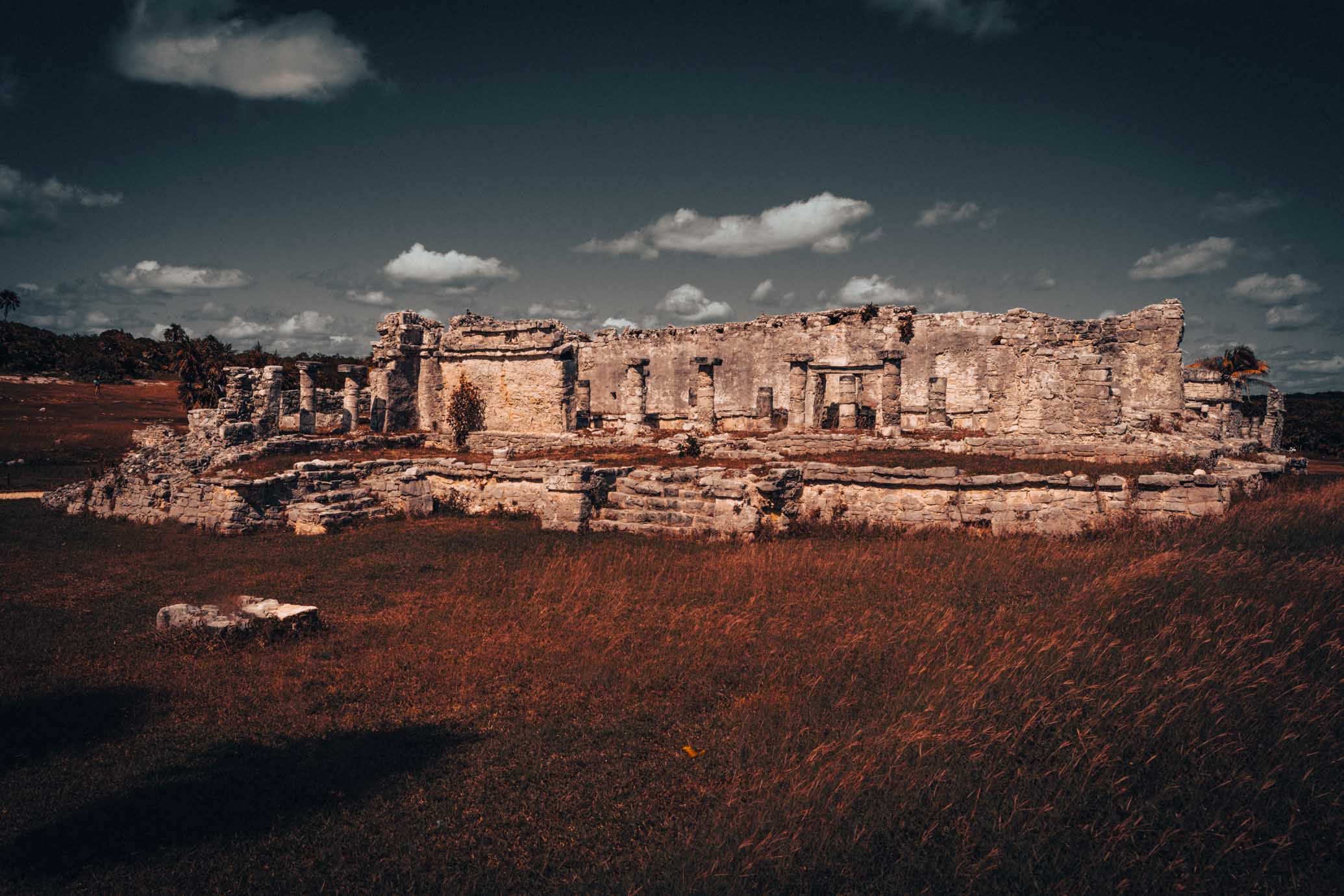 Wide view of ancient Maya stone buildings, columns, and archaeological ruins at Tulum beneath a partly cloudy sky.