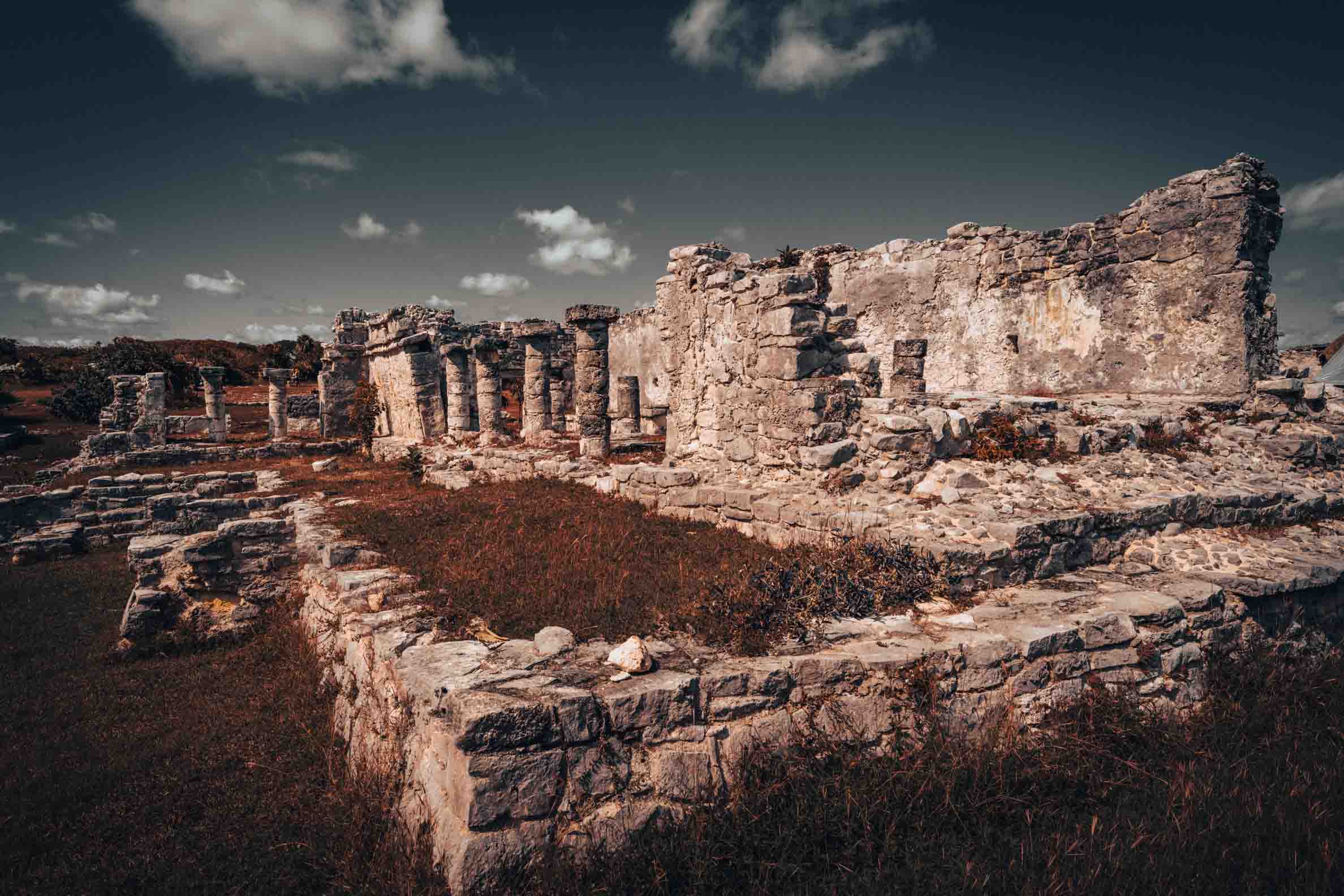 Weathered limestone steps winding through ancient Maya ruins toward a stone temple beneath a cloudy sky at Tulum, Mexico.