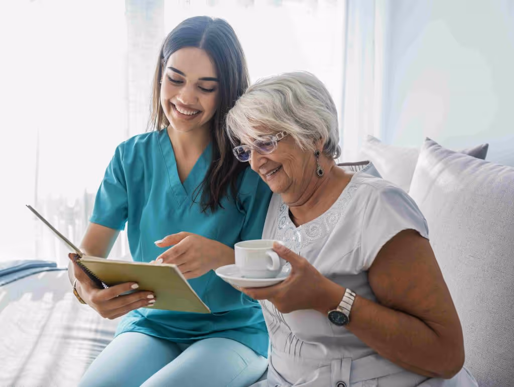 A carer happily reading a notebook to a dementia patient who is holding a cup