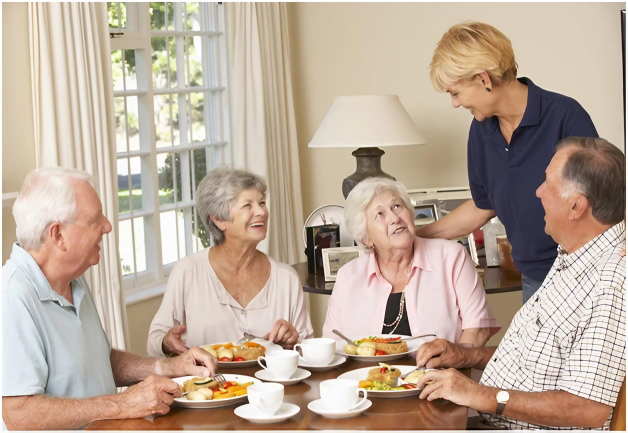 A group of seniors eating together around a round table, and a carer is greeting them