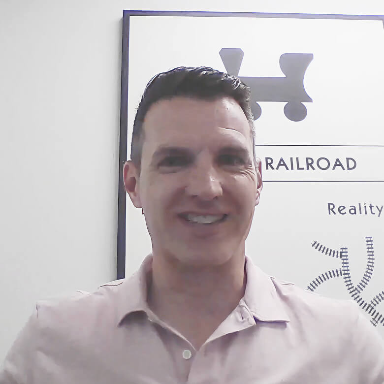 Man smiling in a light pink shirt giving a testimonial for Always Beyond IT services with railroad-themed wall art behind ...