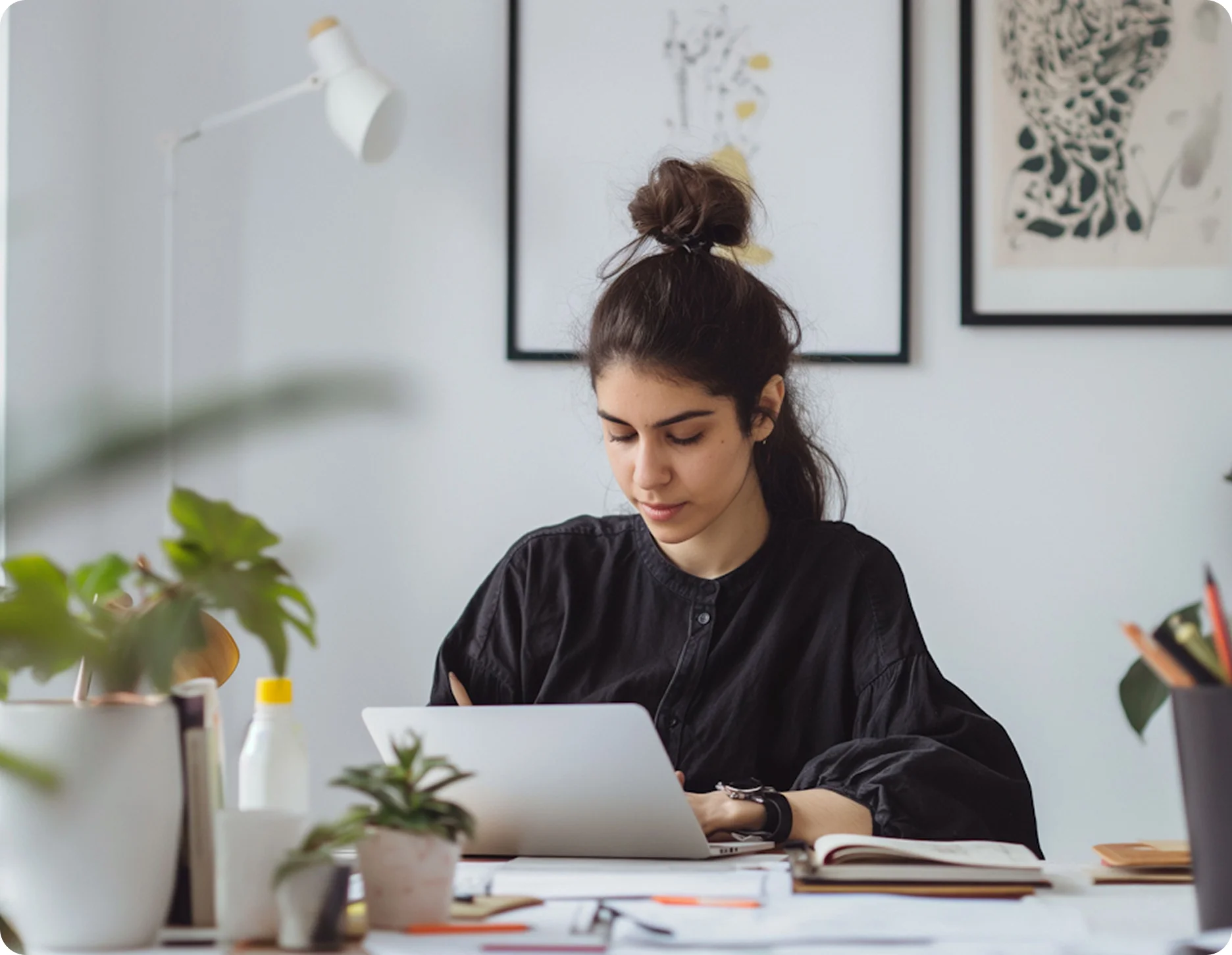 Young woman with hair in a bun working on a laptop at a desk surrounded by plants and office supplies.