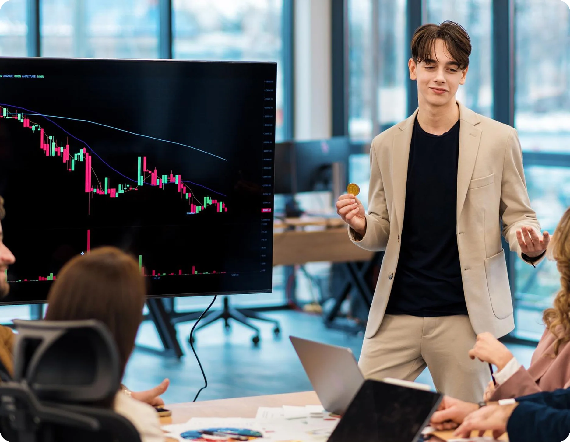Young man in beige suit holding a cryptocurrency coin and presenting in front of a screen with a downward stock chart to colleagues in an office.