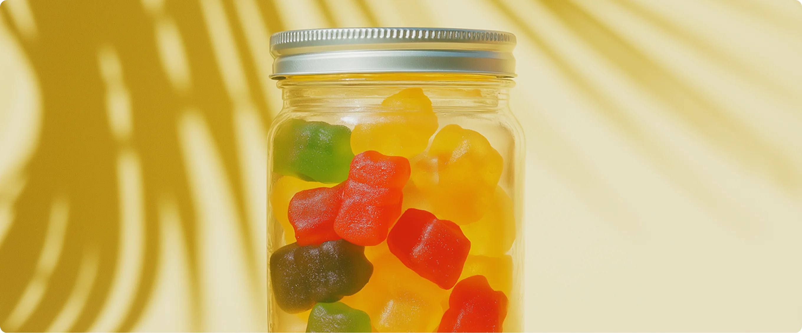 Glass jar filled with colorful gummy bears against a yellow background with shadows.