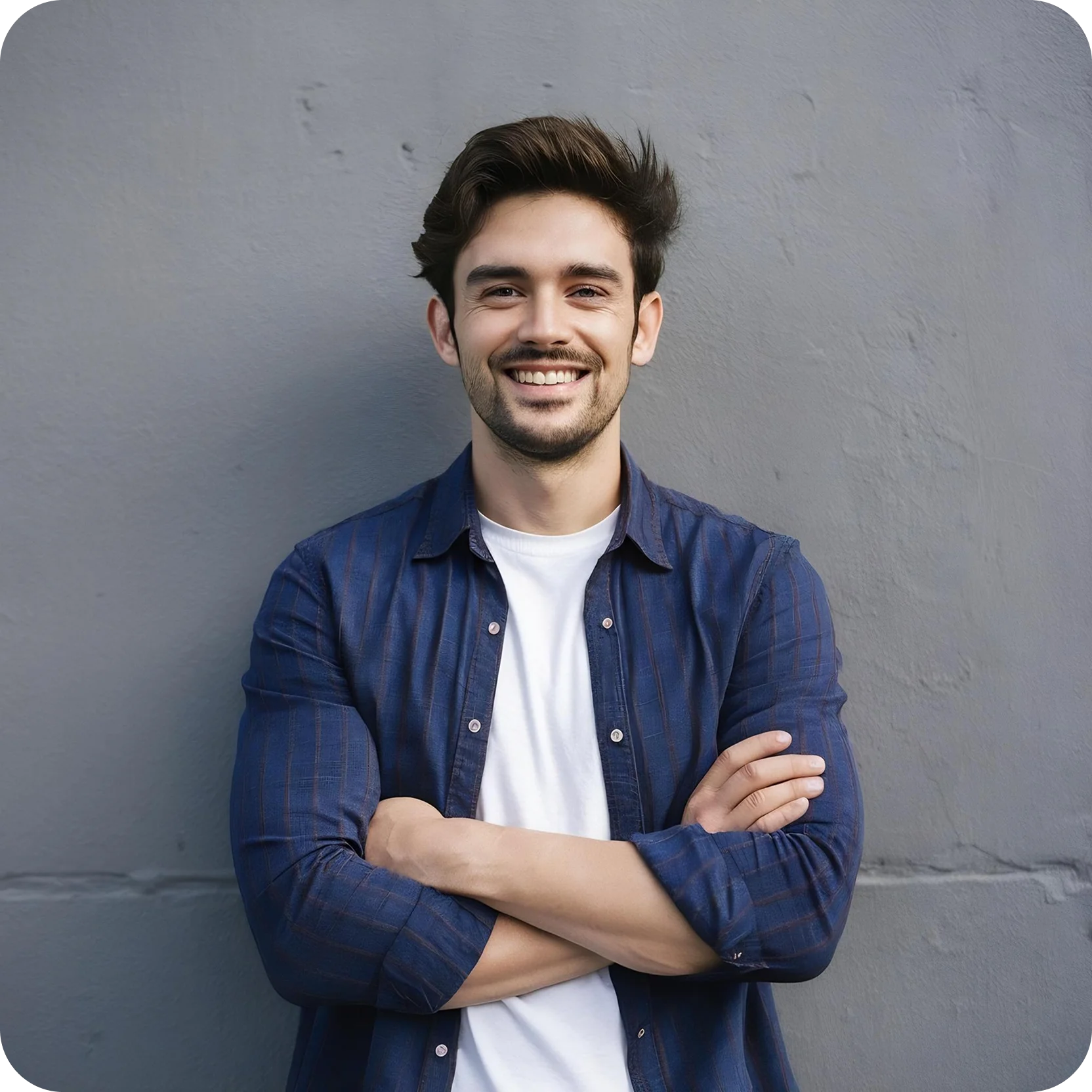 Smiling young man with short dark hair, wearing a blue shirt over a white t-shirt, standing with arms crossed against a gray wall.