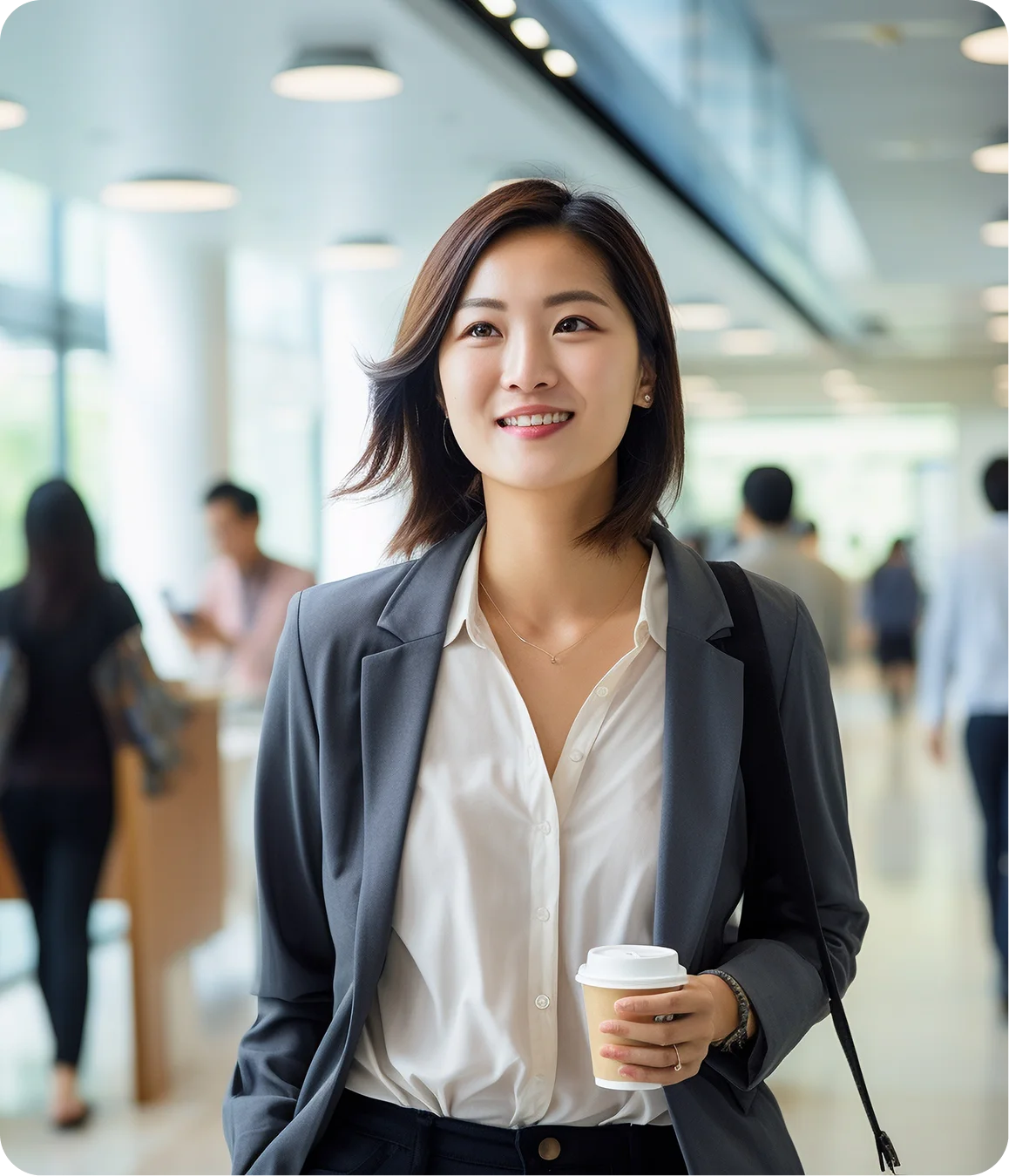 Smiling young businesswoman wearing a gray blazer and white shirt, holding a takeaway coffee cup in an office hallway.
