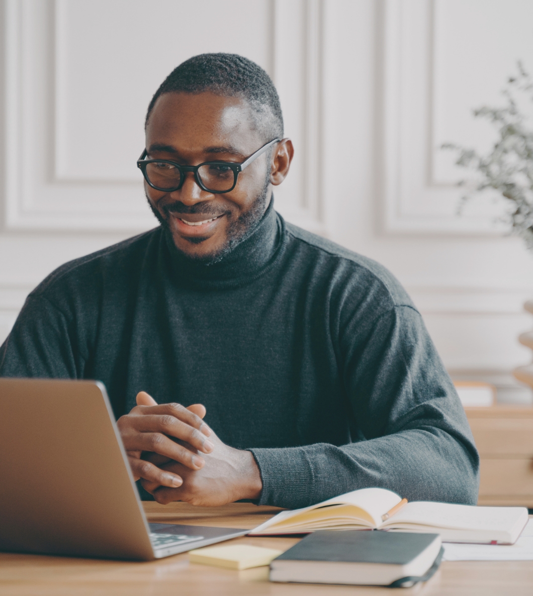 Smiling man with glasses in a dark turtleneck sitting at a desk with a laptop and books.