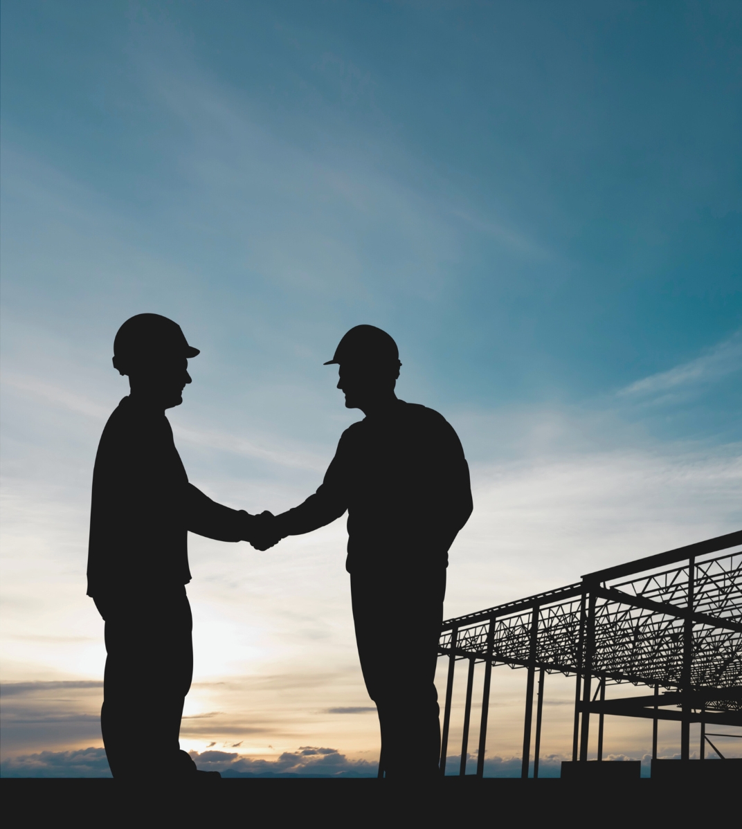 Silhouettes of two construction workers shaking hands by a building framework at sunset.