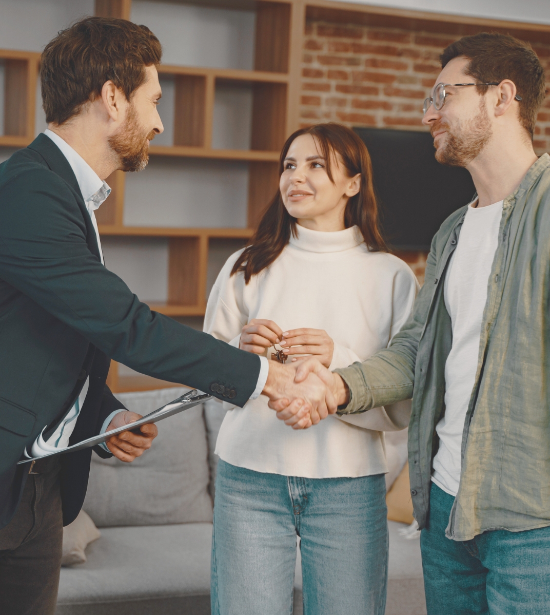 Man in blazer shaking hands with another man while woman watches holding keys indoors.