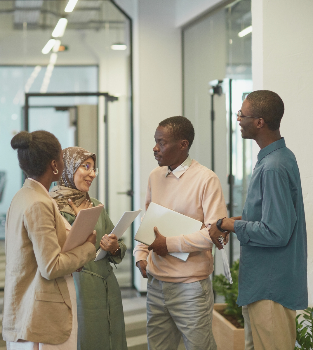 Four diverse professionals holding documents and laptop, engaged in conversation in modern office.