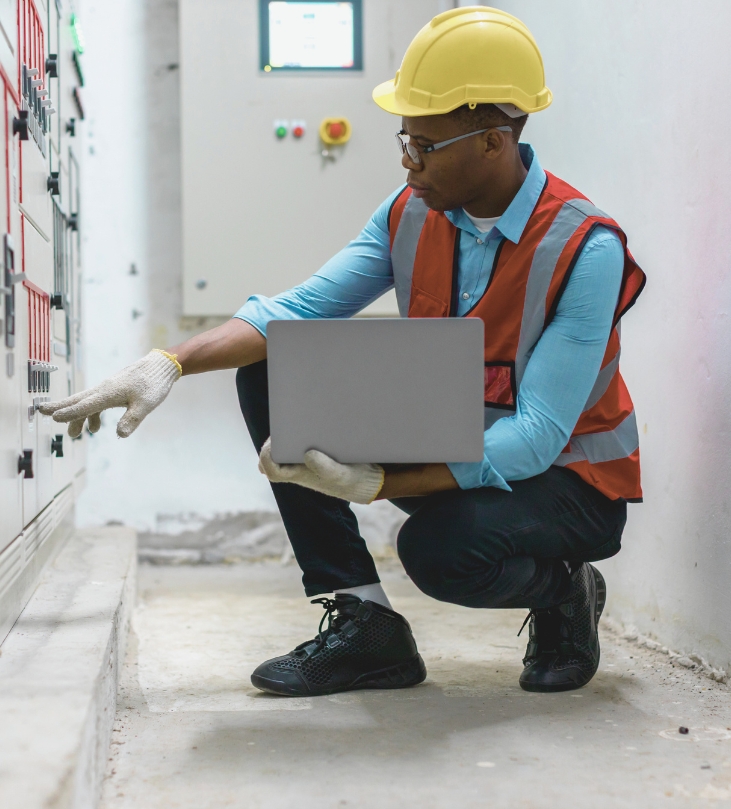 Technician in hard hat and vest holding laptop, adjusting controls on electrical panel indoors.