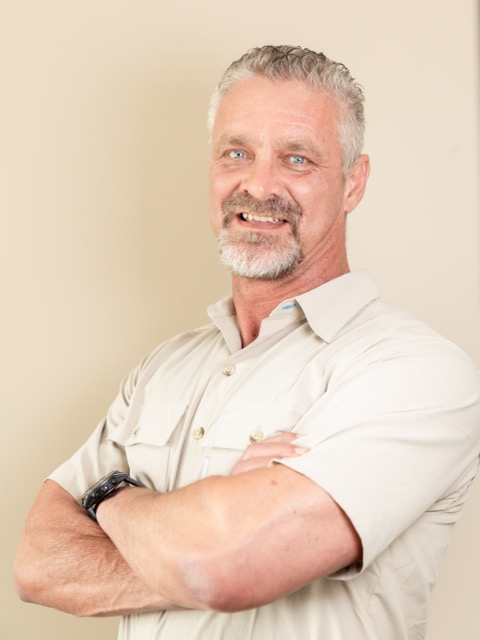 Middle-aged man with gray hair and beard smiling with arms crossed, wearing a beige shirt and watch.