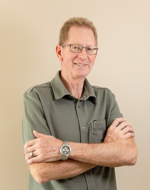 Smiling older man wearing glasses and a green button-up shirt with folded arms.
