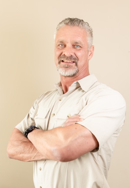 Confident middle-aged man with gray hair and beard, wearing light shirt, arms crossed, smiling.