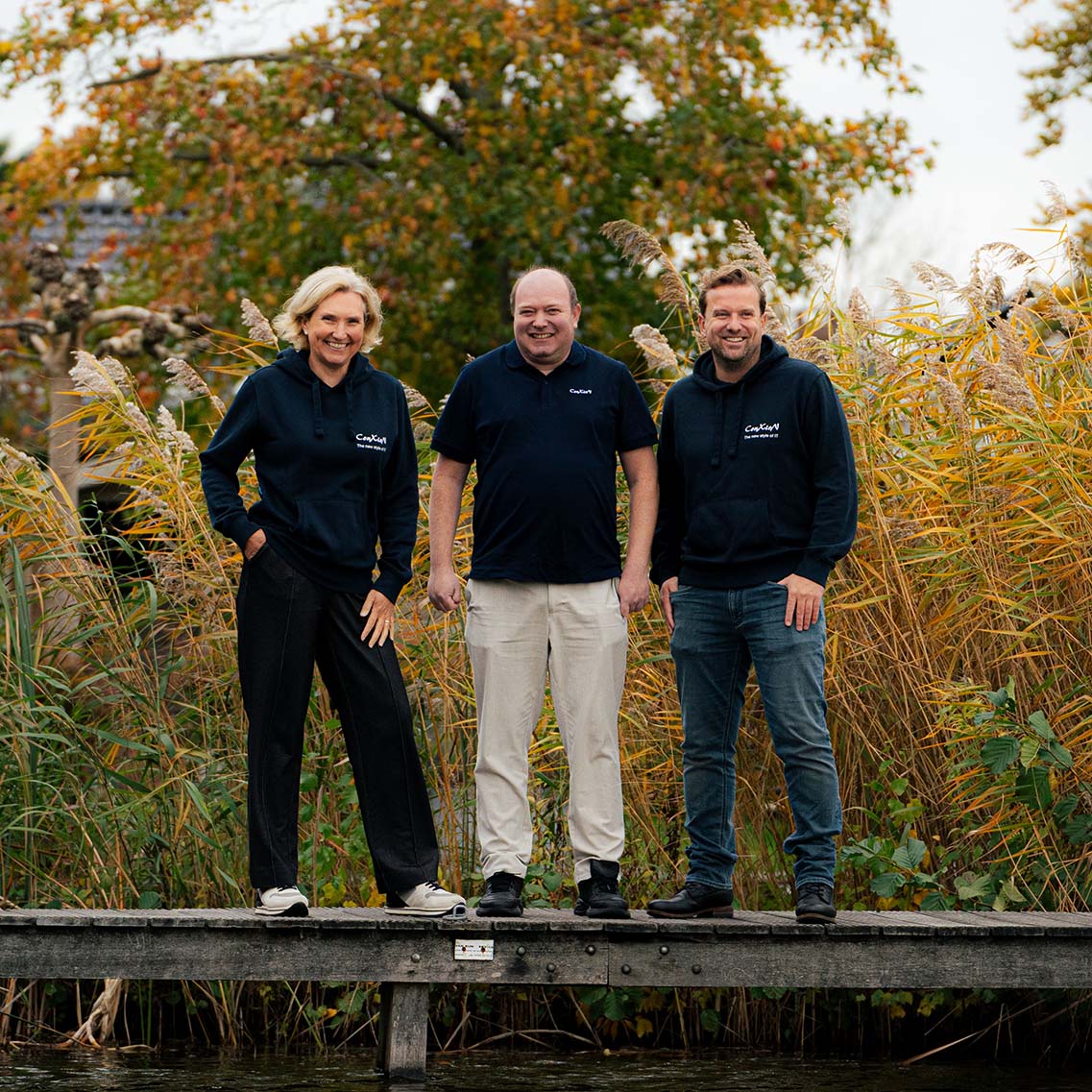 Drie ConXioN-collega’s poseren buiten op een houten brug, met riet en herfstkleuren op de achtergrond.