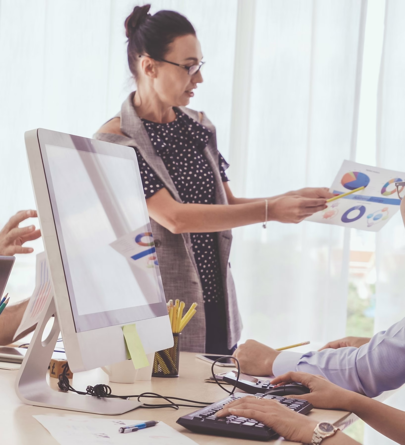Team in einem modernen Büro bei einer Besprechung – eine Frau präsentiert Diagramme auf Papier, während Kollegen am Tisch mit Laptops und Monitoren arbeiten und aufmerksam zuhören.