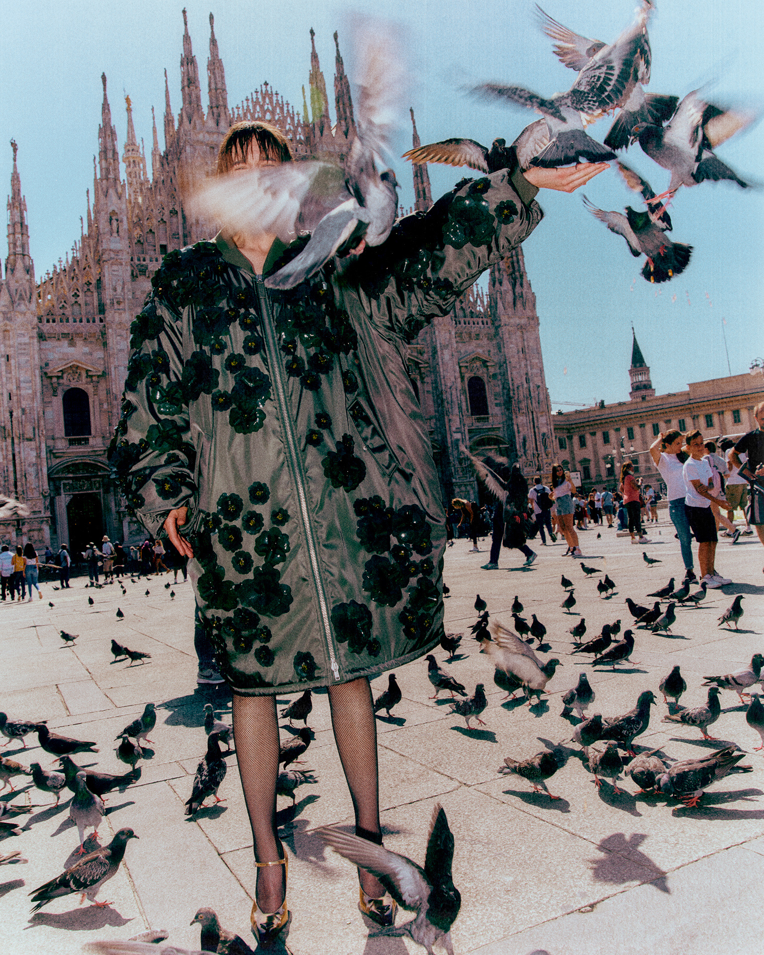 Model in an embroidered green coat in front of Milan Cathedral, pigeons flying up around her in bright sun