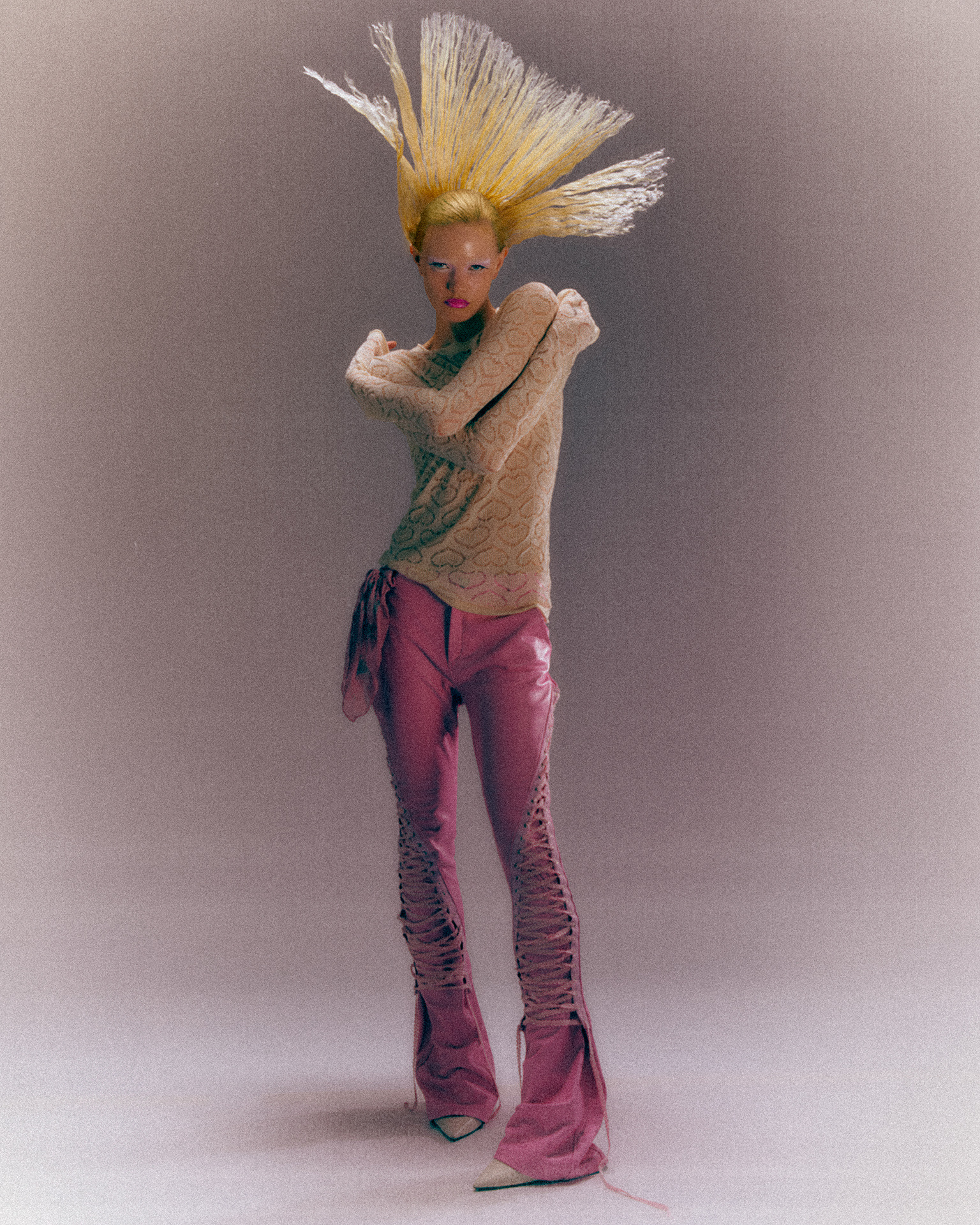 Full-length portrait of a model with stiff blonde hair standing up, in a lace knit and pink trousers, in warm, matte colour