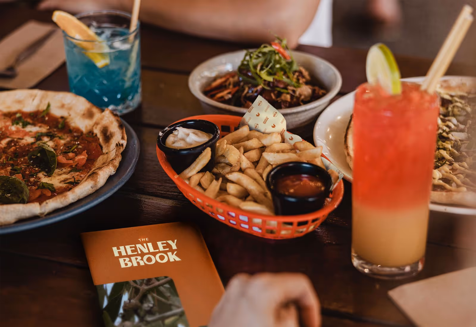 Close-up of a table at The Henley Brook with pizza, fries, cocktails, and branded menu