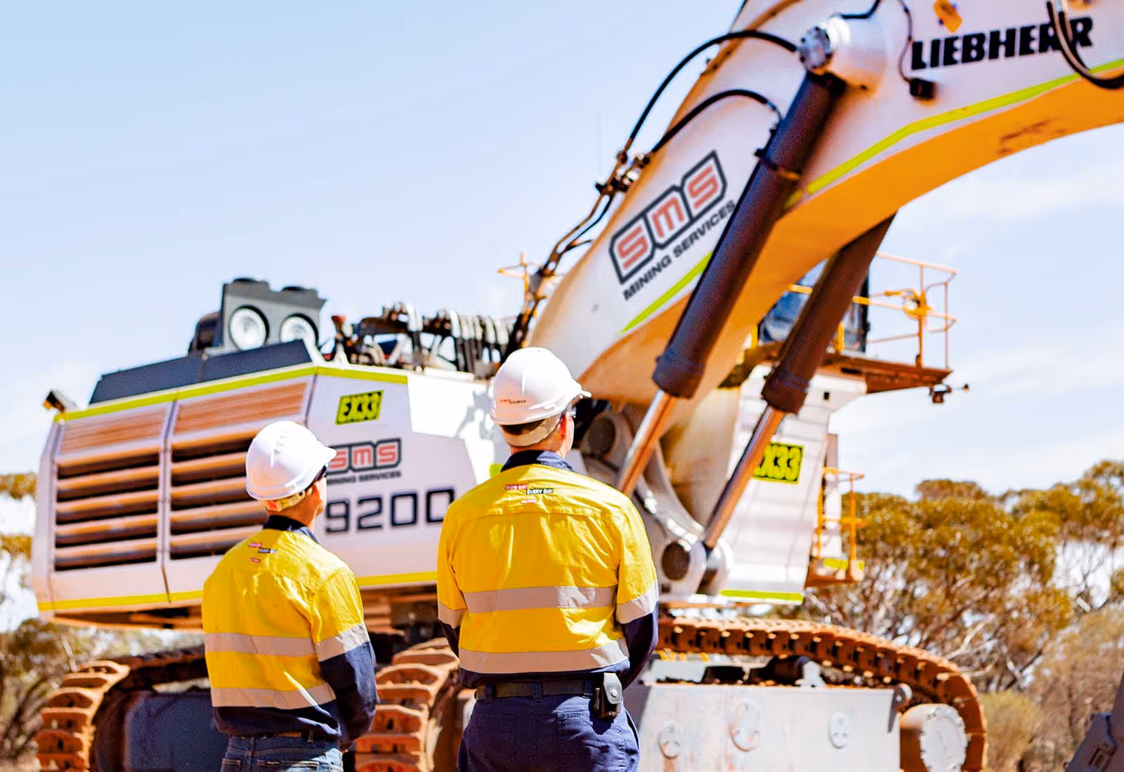 Two workers in yellow hi-vis uniforms observing an SMS-branded Liebherr excavator on a mining site.