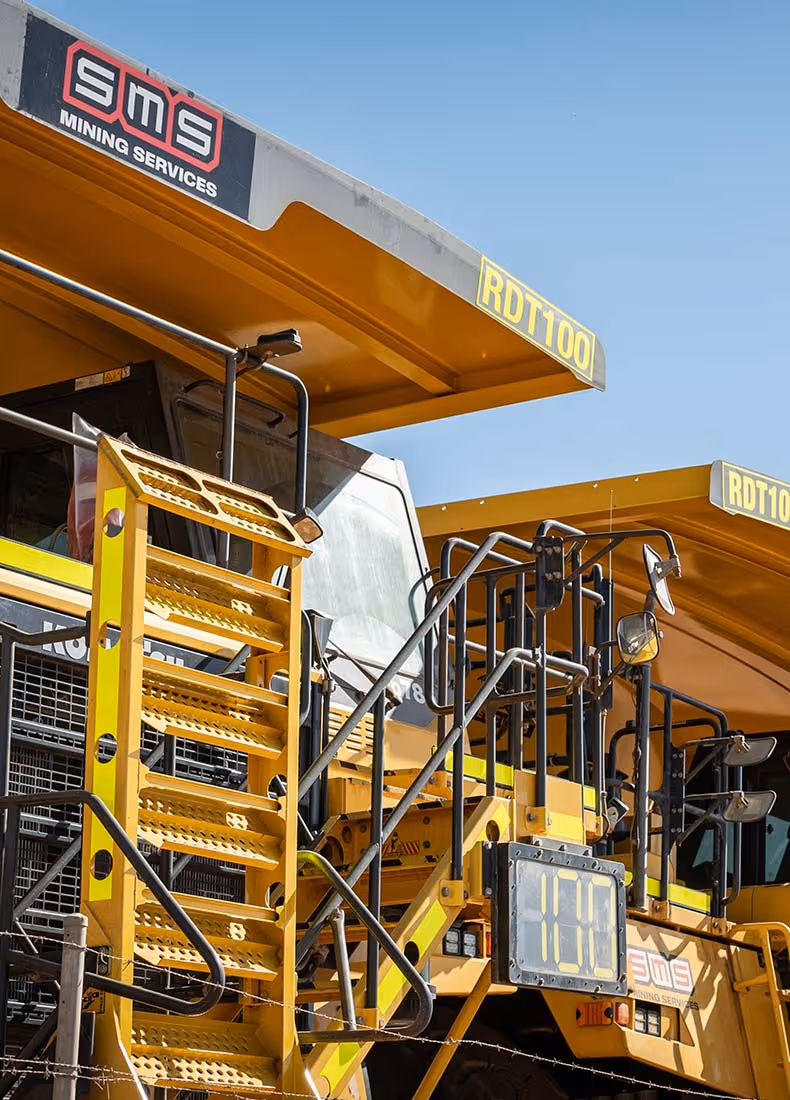 Yellow mining haul trucks with SMS Mining Services branding parked under a clear blue sky.