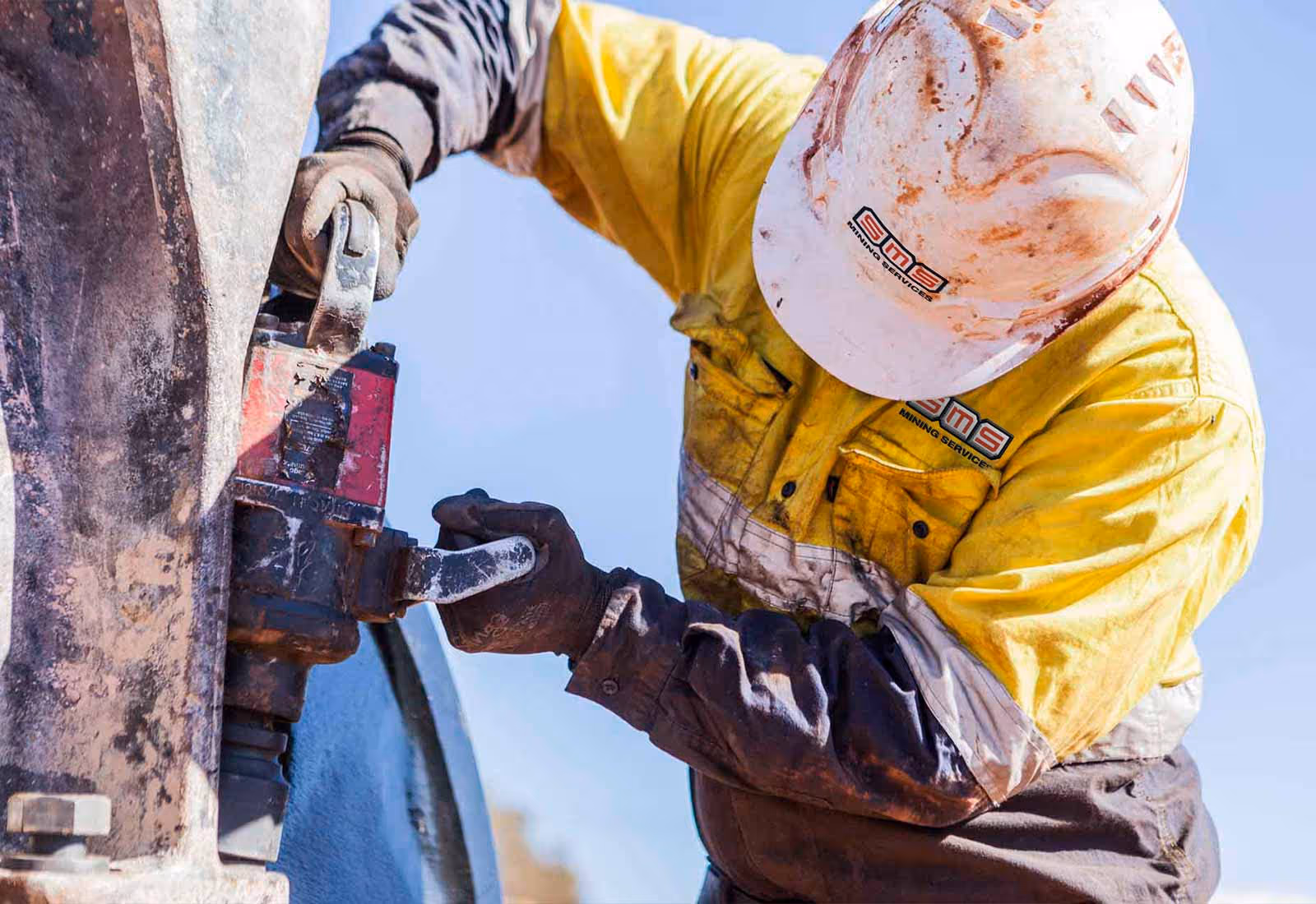 Worker in a yellow SMS-branded shirt and helmet performing maintenance on mining equipment in a dusty environment.