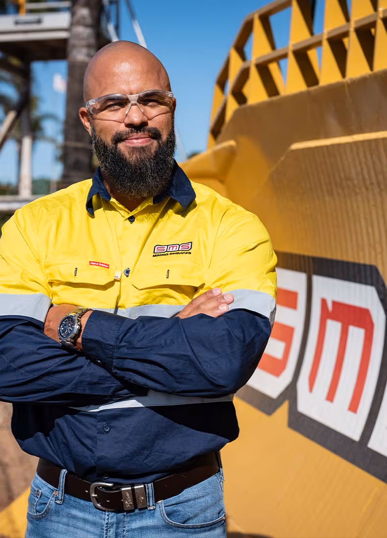 Close-up of SMS Mining Services worker in yellow safety shirt standing confidently in front of branded equipment.