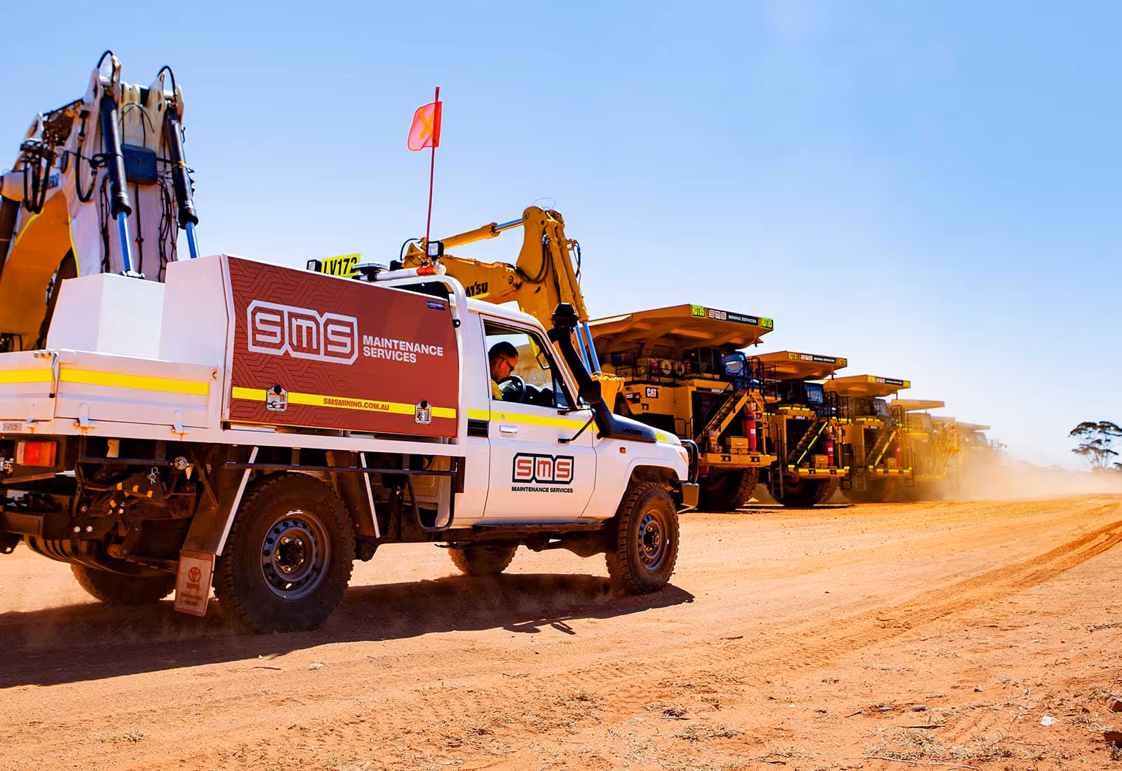 SMS Maintenance Services field vehicle driving on a dirt site with large mining trucks in the background.