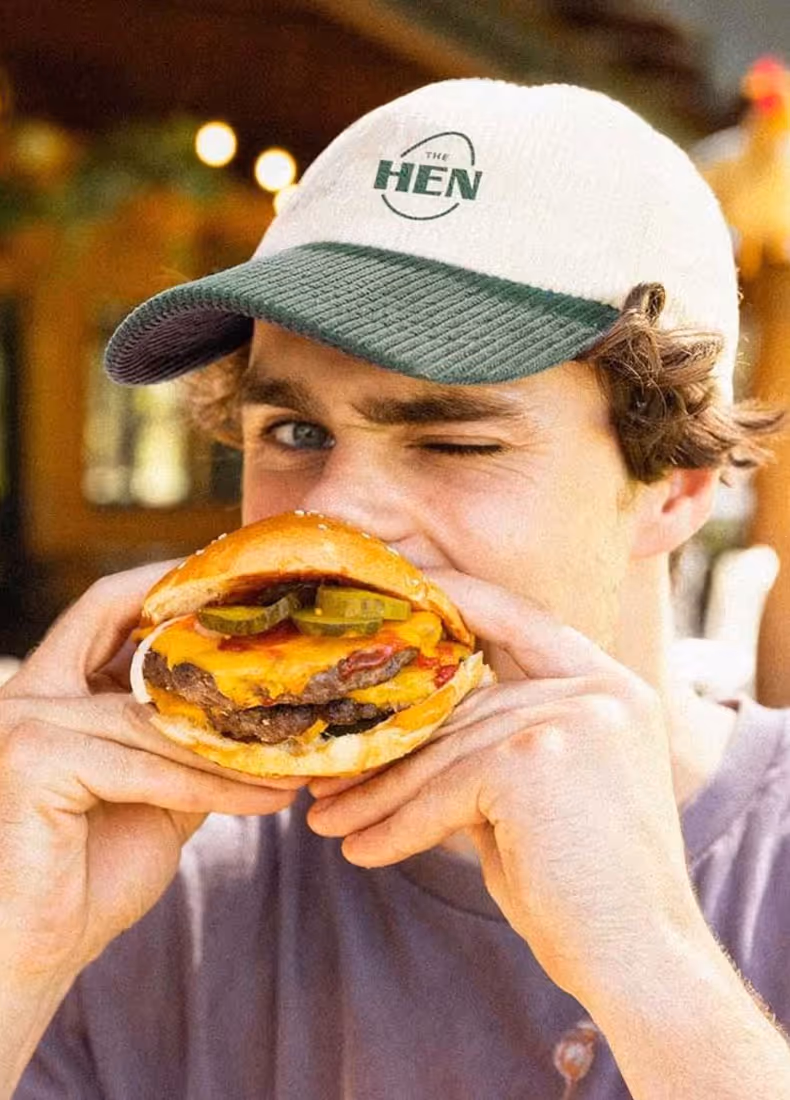 Man wearing a white and green cap with "The Hen" logo, holding and about to bite into a cheeseburger with pickles and bacon, smiling with one eye closed.