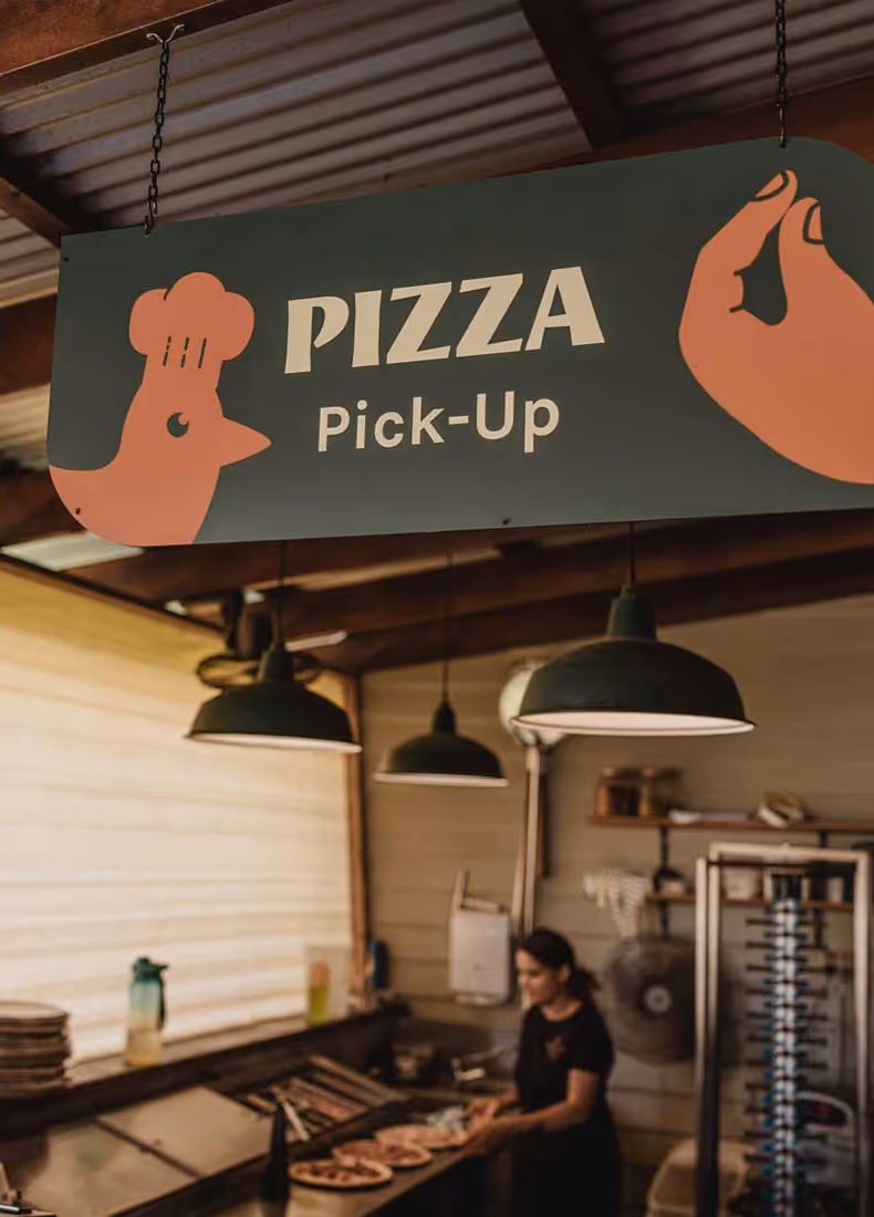 Green hanging sign reading “Pizza Pick-Up” with cartoon hands and chef illustration, inside a rustic kitchen with staff preparing pizzas.
