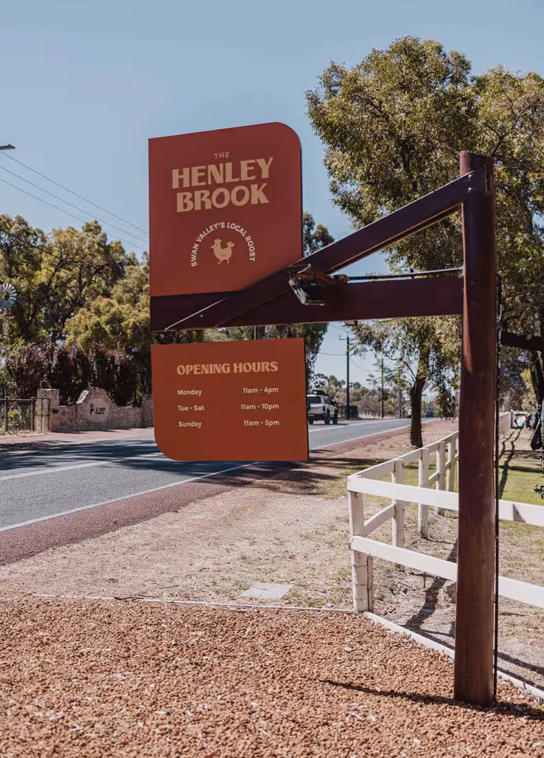 Large roadside sign for The Henley Brook showing opening hours, set against a sunny day with trees and a white fence along the road.