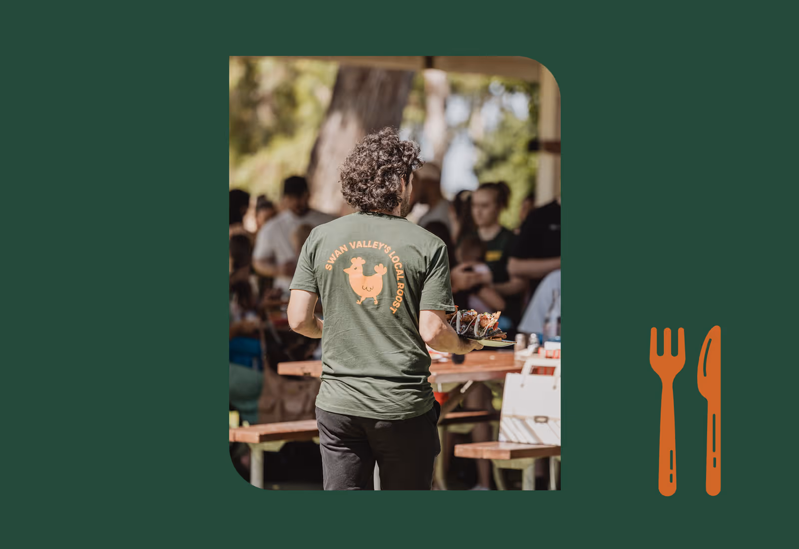Waiter seen from behind wearing a green “Swan Valley’s Local Roost” t-shirt, carrying plates of food at an outdoor dining area.