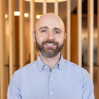 Team photo of a smiling man with a beard, in a button-up shirt, standing confidently against a modern wood-panel backdrop—featured in Four Stripes' team section.