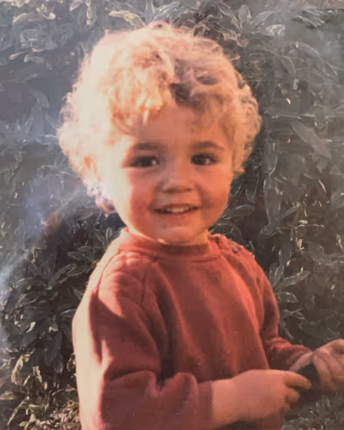 Vintage childhood photo of a curly-haired toddler in a red sweater, smiling at the camera in a leafy backyard setting—part of the fun and personal team series at Four Stripes.
