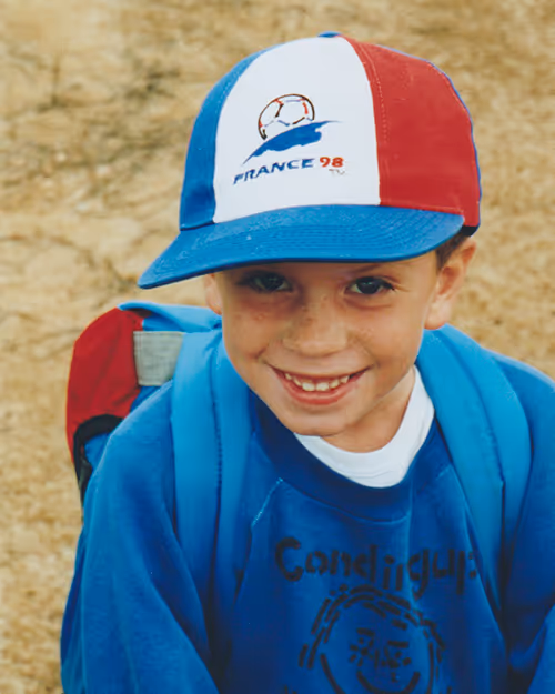 Childhood photo of a smiling boy in a France 98 cap and blue backpack, standing outdoors on a dirt path—highlighting a nostalgic team profile at Four Stripes.