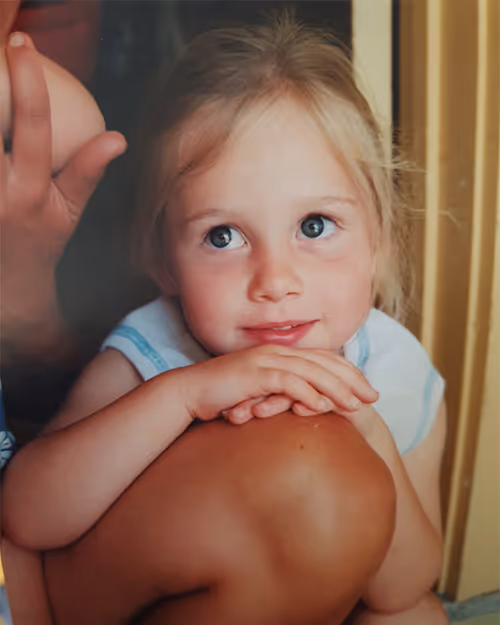 Young girl with bright blue eyes and blonde hair, resting her hands on her knees and looking up thoughtfully—used for a personal touch in Four Stripes team profiles.
