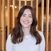 Team photo of a smiling woman in a white ribbed top with long brown hair, posed against a modern timber wall—part of the Four Stripes team profile gallery.
