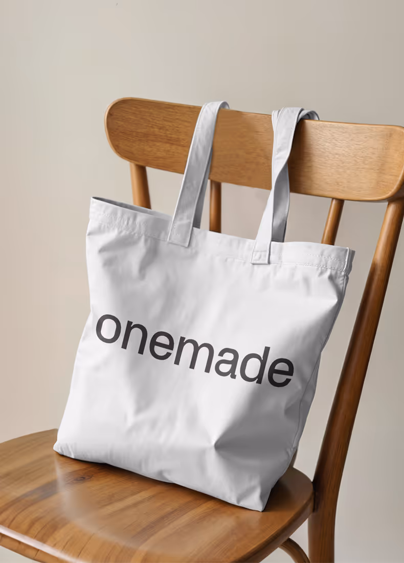White tote bag with bold black Onemade logo draped over wooden chair.