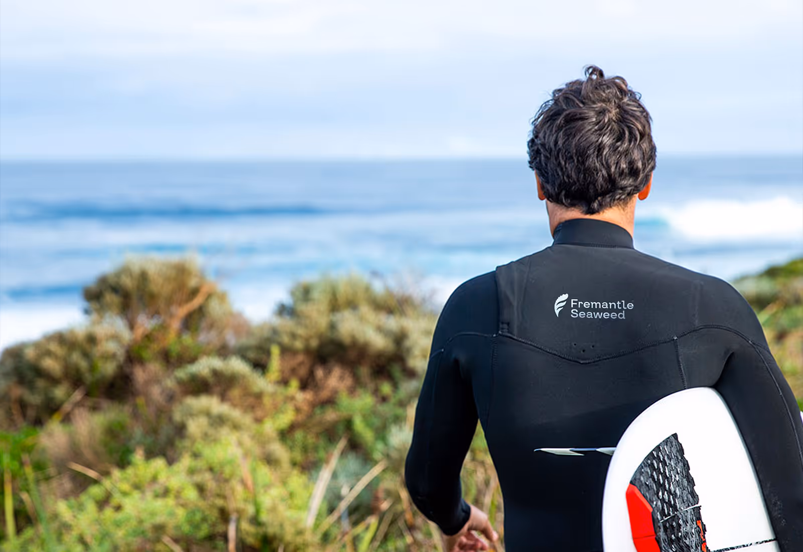 Surfer in wetsuit with Fremantle Seaweed logo holding surfboard and looking at ocean.