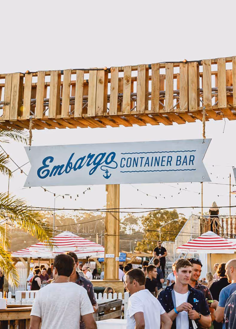 Outdoor wooden structure with a hanging sign reading “Embargo Container Bar” in blue lettering with an anchor icon, above a crowd of people enjoying a lively outdoor gathering under string lights and striped umbrellas at sunset.