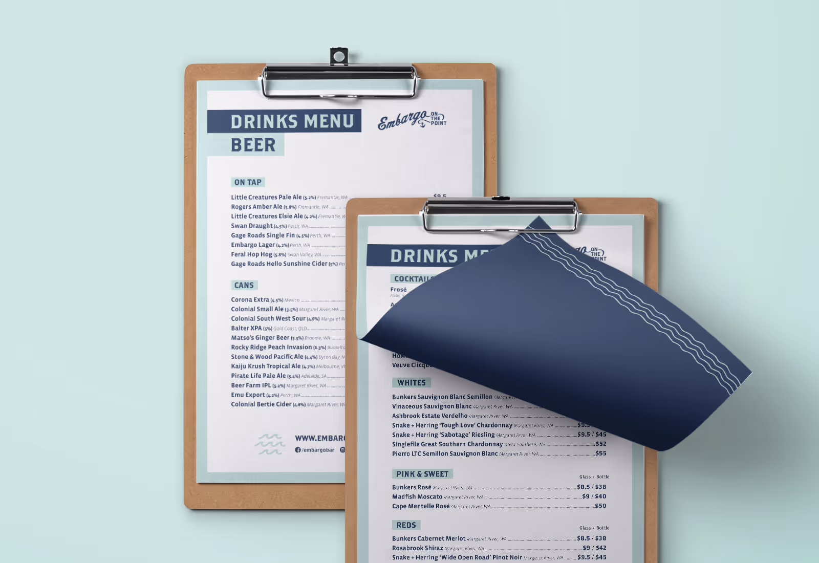 Close-up of two drinks menus clipped to wooden boards, featuring an extensive selection of beers, cocktails, and wines for Embargo on the Point, with a minimalist nautical-themed design in navy and aqua.