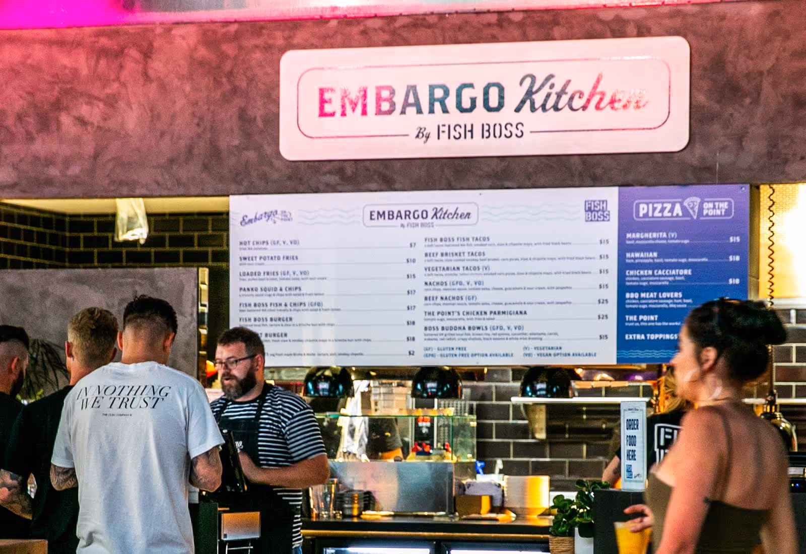 Indoor bar counter scene at Embargo Kitchen by Fish Boss, with staff preparing orders under warm lighting, a large menu board displaying a range of meals including fish tacos, burgers, pizzas, and fries, and customers ordering at the counter.