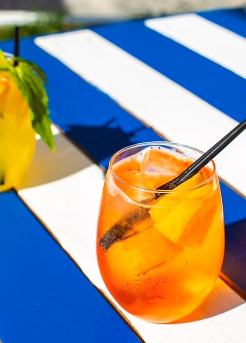 Refreshing orange cocktail served in a rounded glass with ice and fruit garnish, a black straw, and condensation on the glass, placed on a sunlit blue and white striped outdoor table.