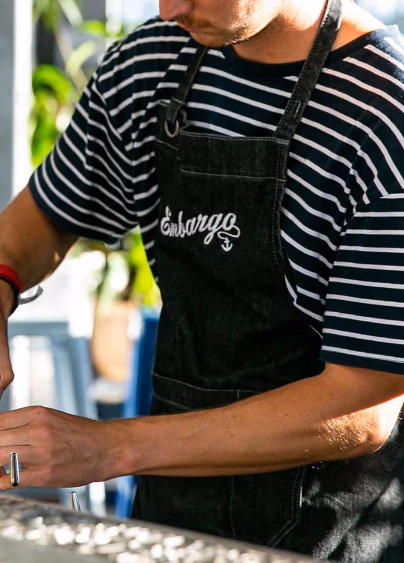 Bartender wearing a navy-and-white striped shirt and a dark apron embroidered with the “Embargo” logo and anchor icon, preparing a drink at the bar with sunlight casting across their hands.