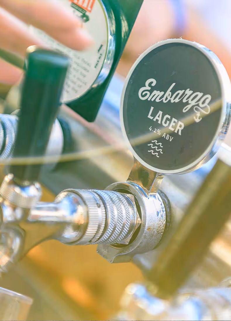 Close-up of a beer tap with the Embargo Lager branding, displaying a sleek black-and-white label with an anchor icon and “4.2% ABV,” set against a softly blurred bar background.