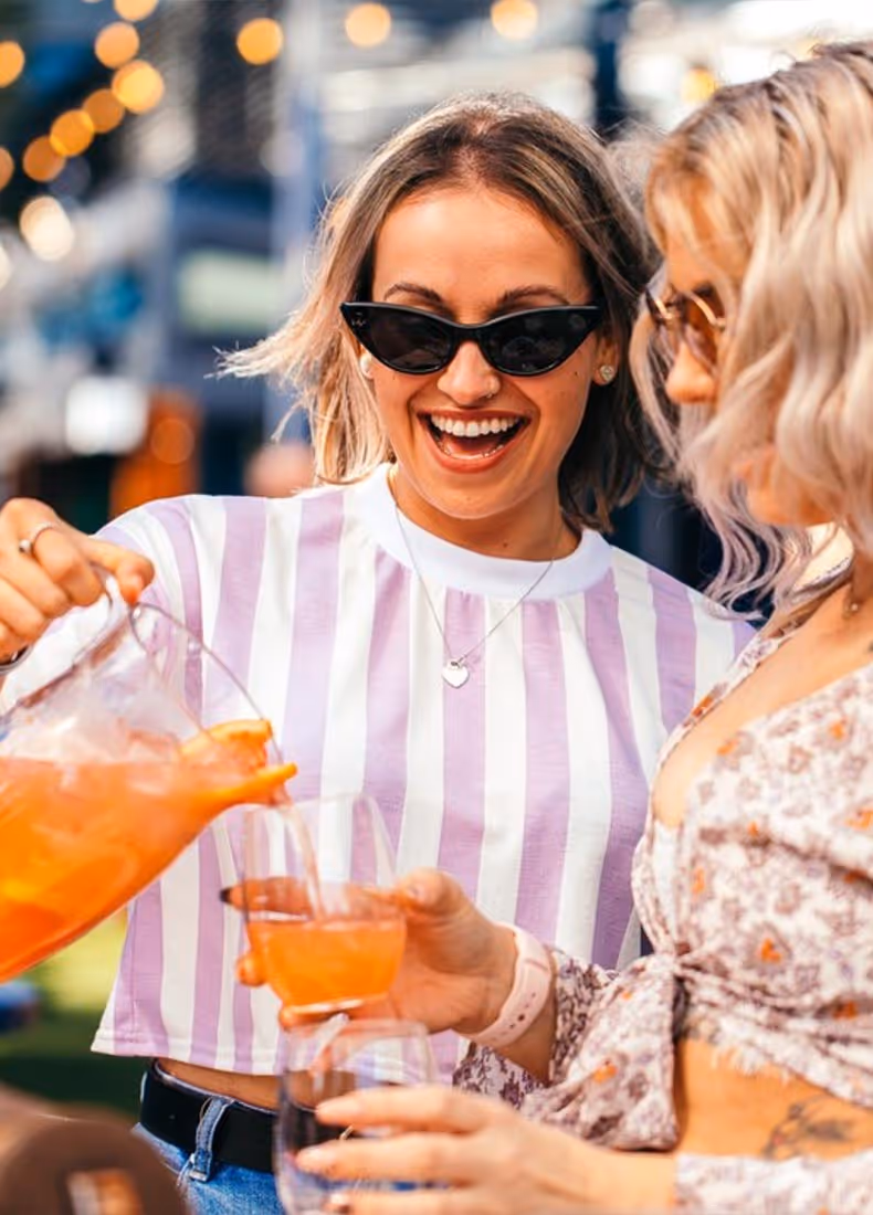 Two women at Embargo On The Point smiling and enjoying a drink service moment, with one pouring a vibrant orange cocktail from a jug into stemless glasses, dressed in summery striped and floral outfits.