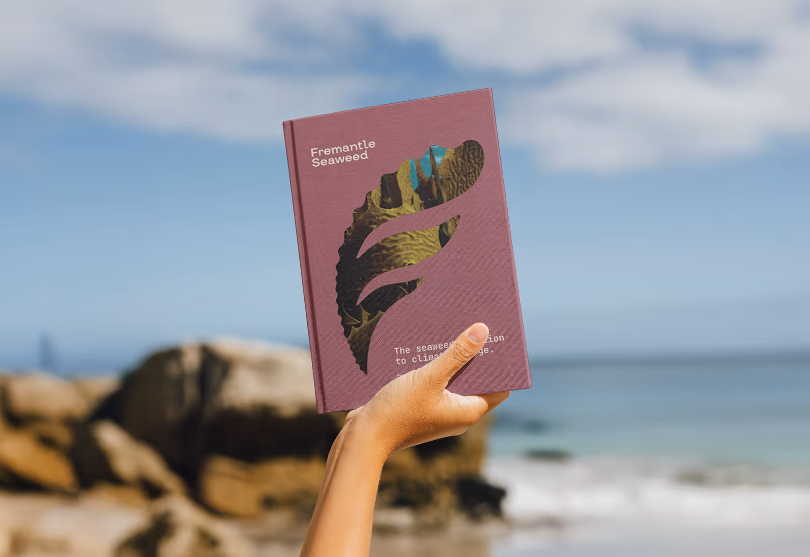 Hand holding a Fremantle Seaweed branded hardcover book against a coastal backdrop, featuring a mauve cover with a seaweed-shaped cutout and the tagline 'The seaweed solution to climate change.'