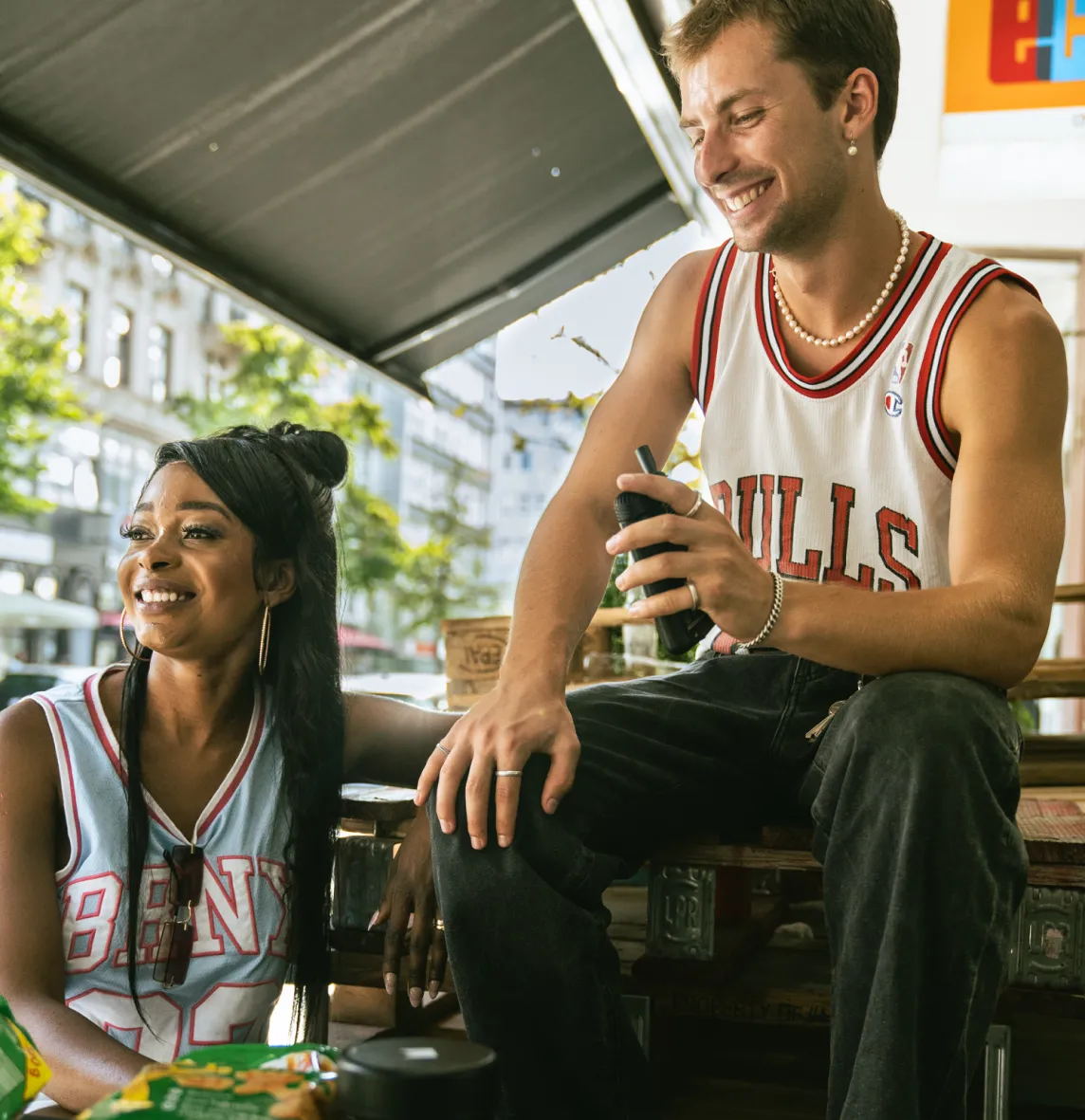 Lächelnder Mann und Frau sitzen und unterhalten sich im Freien, vor ihnen auf dem Tisch stehen Snack-Tüten.