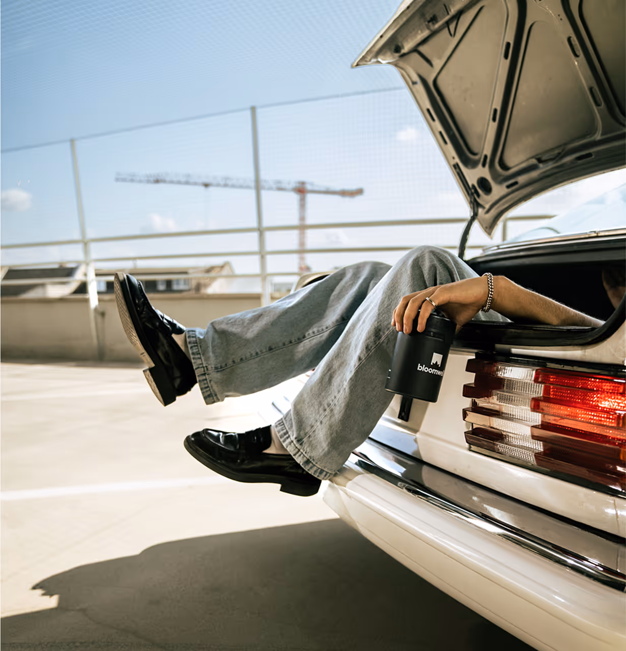 Person in Jeans und schwarzen Schuhen, die mit den Beinen aus einem offenen Kofferraum heraushängt und einen schwarzen Becher in der Hand hält.