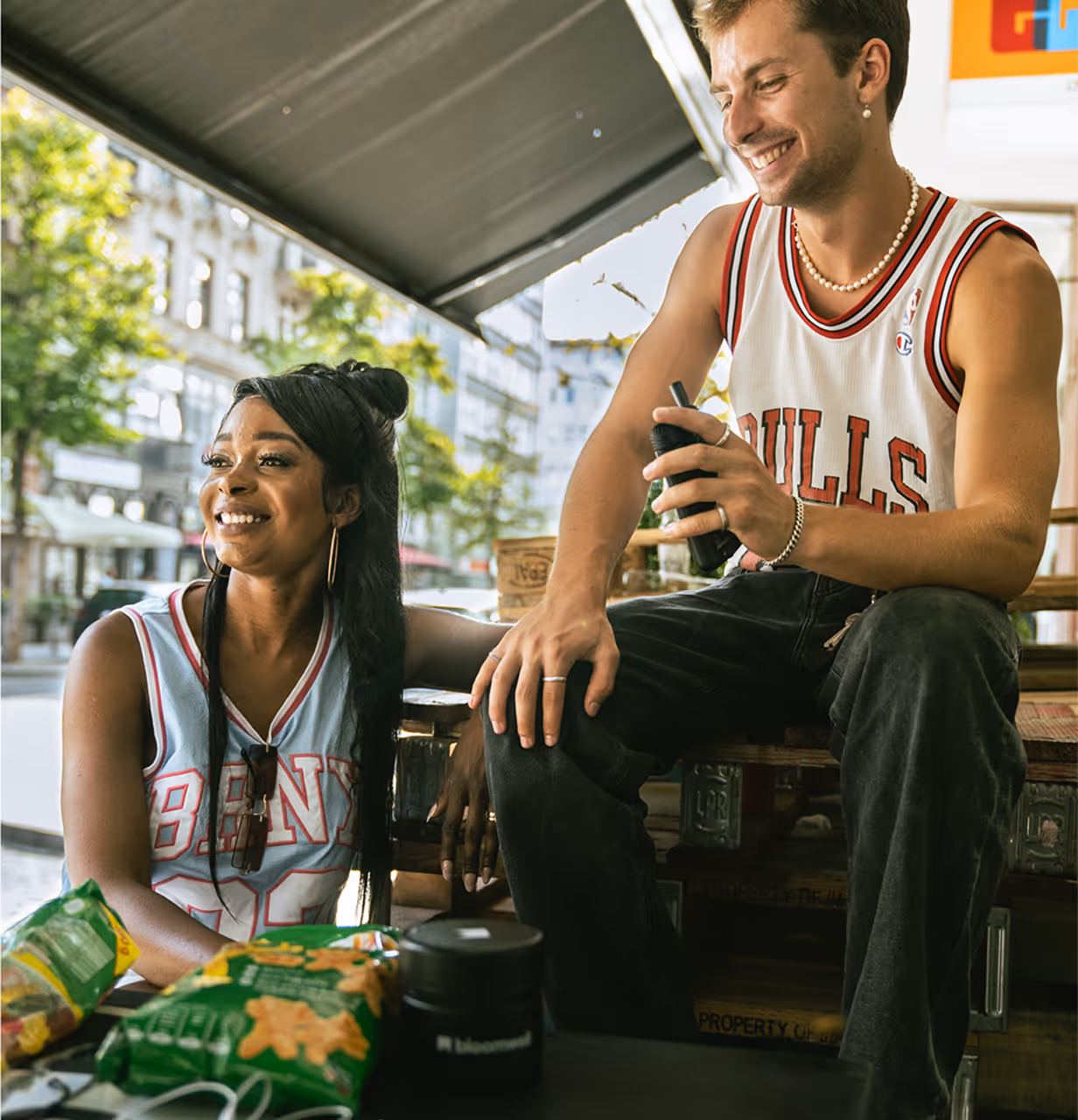 Lächelnder Mann und Frau sitzen und unterhalten sich im Freien, vor ihnen auf dem Tisch stehen Snack-Tüten.